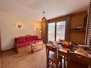 A cozy room with a wooden sofa, a dining table with chairs, and a view of a mountainous landscape through the window. - Apartment mountain view - 4 guests (Vars)
