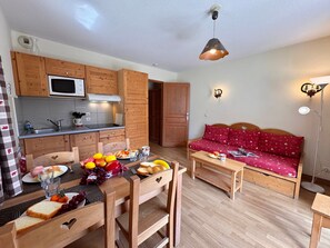 Un espace de vie compact comprenant un coin cuisine, un coin repas avec une table pour deux personnes et un canapé recouvert d’un tissu à motifs en forme de cœur.