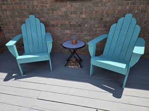 Two Adirondack chairs with a small round table and a potted plant between them, set on a wooden deck against a brick wall.