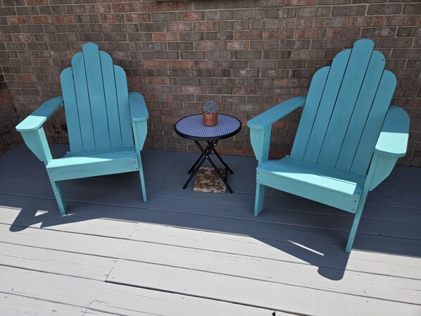 Two Adirondack chairs with a small round table and a potted plant between them, set on a wooden deck against a brick wall.