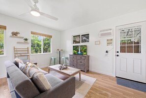 A living room with a sofa, coffee table, and a ceiling fan. There are windows with views of greenery and a door with a windowed section. - Nani Mala Cove at Island Rainforest Grove (Keaau)