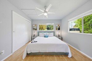 A bedroom with a bed, a ceiling fan, a window with a view of greenery, and artwork on the wall. - Malie Kai House at Island Rainforest Grove (Keaau)