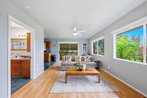 A spacious living area with a sofa, coffee table, and large windows offering a view of greenery. - Malie Kai House at Island Rainforest Grove (Keaau)