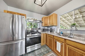A modern kitchen with stainless steel appliances, wooden cabinets, and a large window providing natural light. - Malie Kai House at Island Rainforest Grove (Keaau)