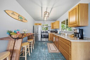 A kitchen with wooden cabinets, a stainless steel refrigerator, an oven, and a microwave. There is a wooden table with chairs and a surfboard decoration on the wall. - Malie Kai House at Island Rainforest Grove (Keaau)