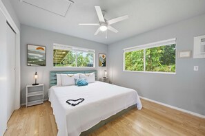 A bedroom with a bed, two bedside tables, a ceiling fan, and two windows. - Malie Kai House at Island Rainforest Grove (Keaau)
