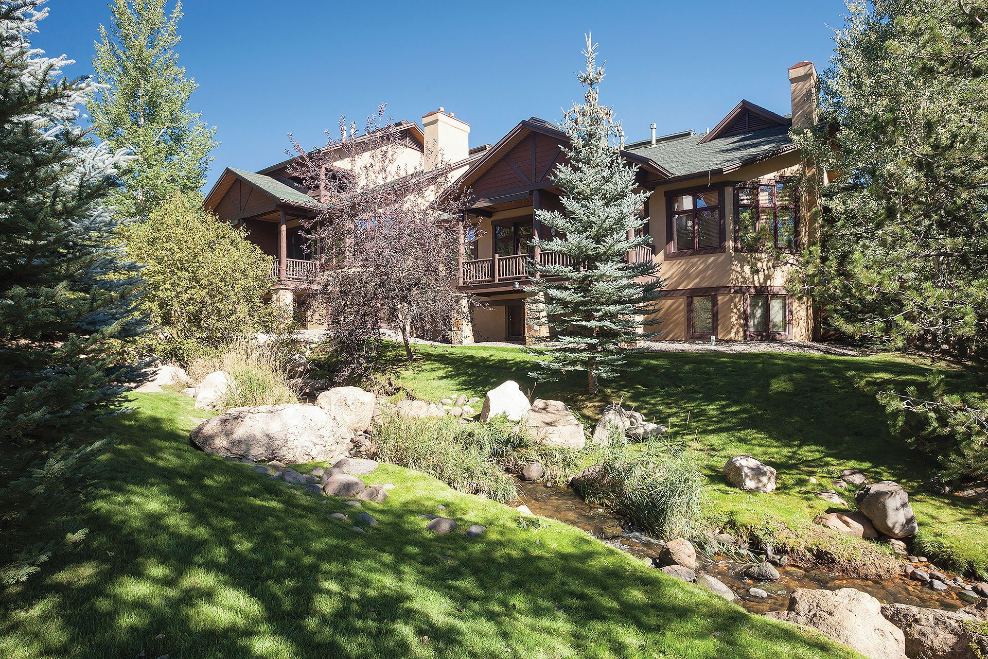 A two-story lodge with a stone facade, a wooden balcony, and a small stream in the foreground.