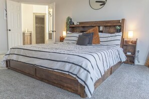 A bedroom with a wooden bed frame, striped bedding, two bedside tables with lamps, and a door leading to a bathroom. - Spacious Arlington 5BR Sleeps 12 Near AT&T Stadium & Stovall Park (Arlington)