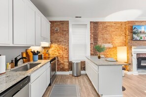 A modern kitchen with white cabinetry, a brick accent wall, a built-in oven, and a small island with a sink. - Historic Soulard 2BR | Stylish Balcony Retreat (St. Louis)