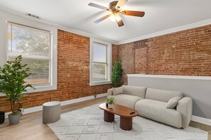 A room with a brick wall, a beige sofa, a wooden coffee table, and a ceiling fan. - Historic Soulard 2BR | Stylish Balcony Retreat (St. Louis)