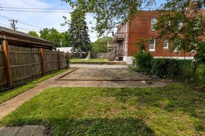 Un jardin à l’arrière d’une maison, avec une clôture en bois, une pelouse verdoyante et un bâtiment en briques en arrière-plan.
