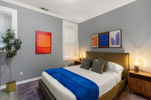 Une chambre moderne avec un grand lit, des lampes de chevet, une plante en pot et des œuvres d’art abstraites accrochées au mur.