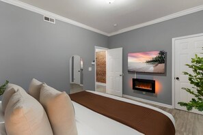 A modern bedroom with a bed, a fireplace, a TV mounted on the wall, and a door leading to another room. - Historic Soulard 2BR | Stylish Balcony Retreat (St. Louis)