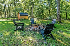 A forested area with a fire pit and three Adirondack chairs.