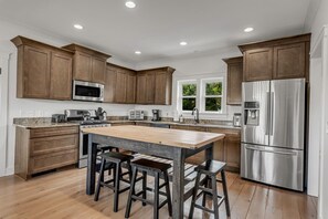 A modern kitchen with wooden cabinets, stainless steel appliances, a central island with stools, and a window providing natural light. - Magical Family Escape: Treehouse, Games, Memories (Spring Hill)