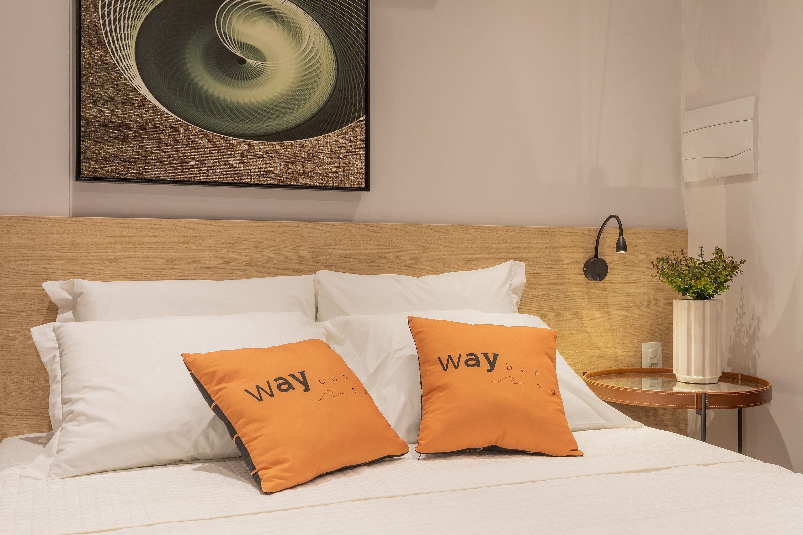 A neatly made bed with white linens, two orange pillows with text, a wooden headboard, a wall-mounted lamp, and a small table with a plant.