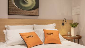 A neatly made bed with white linens, two orange pillows with text, a wooden headboard, a wall-mounted lamp, and a small table with a plant.
