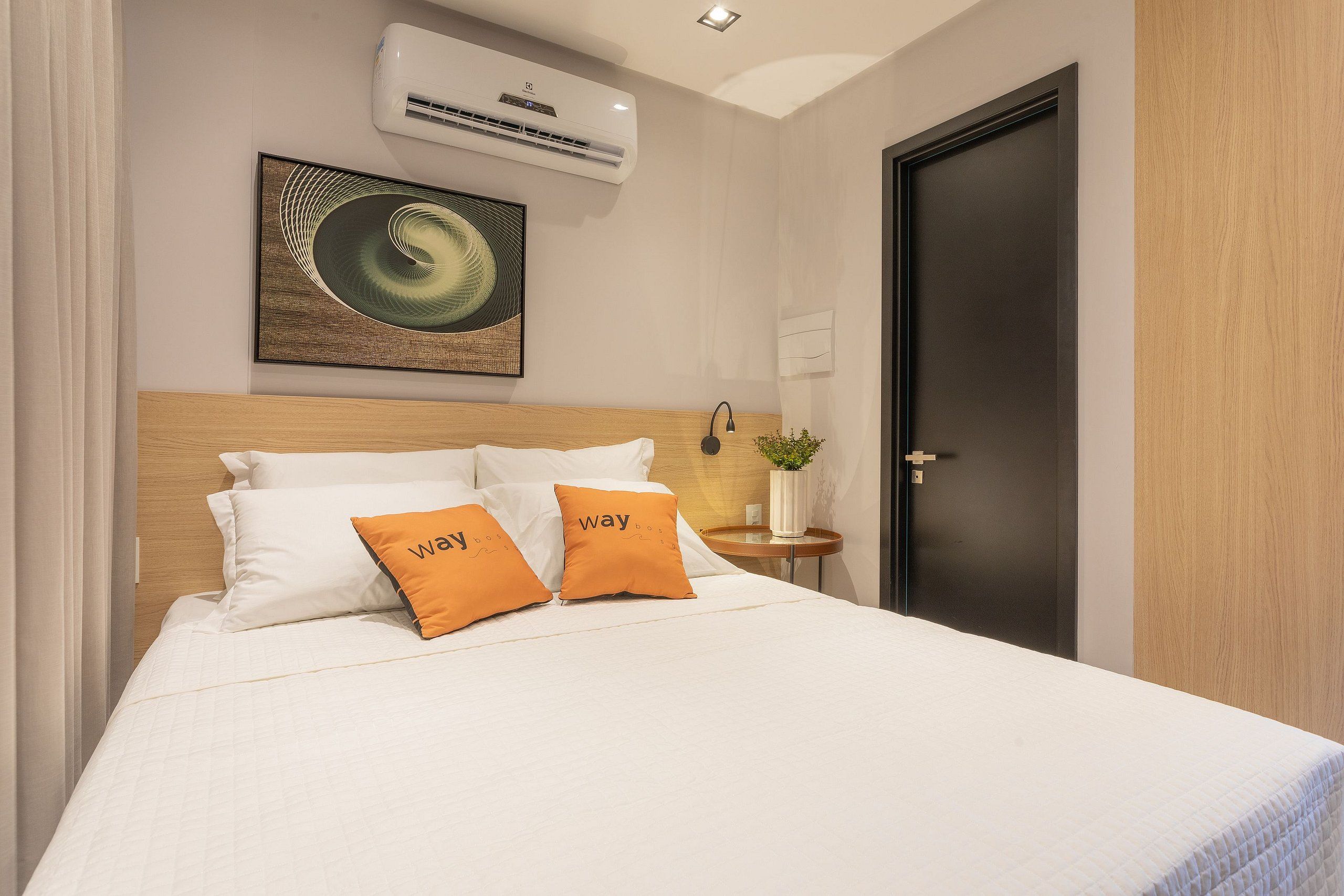A neatly made bed with white and orange pillows, a wall-mounted air conditioner, a framed artwork, and a door with a light switch.