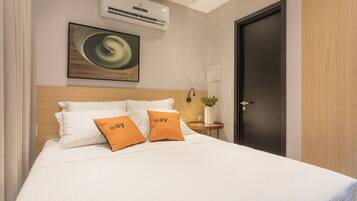 A neatly made bed with white and orange pillows, a wall-mounted air conditioner, a framed artwork, and a door with a light switch.