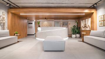 A modern reception area with a reception desk, two sofas, a large ottoman, wooden paneling, and a plant.