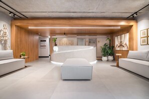 A modern reception area with a reception desk, two sofas, a large ottoman, wooden paneling, and a plant. - Way Bossa #G2 - Flat in Bessa by Carpediem (João Pessoa)