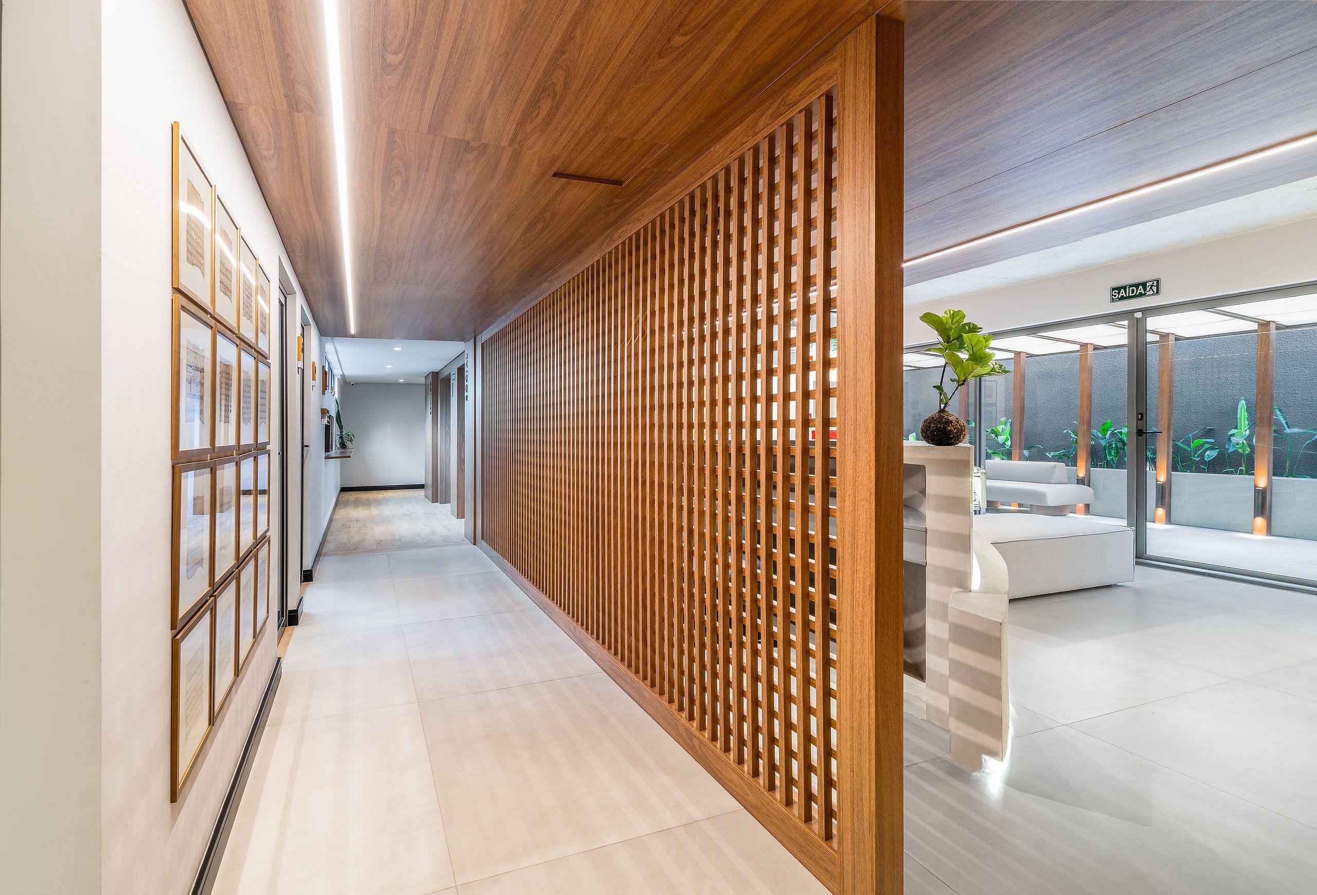Un pasillo con separador de listones de madera, mostrador de recepción y zona de estar con una planta.