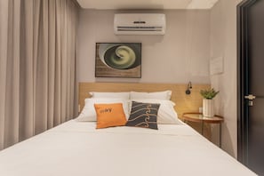 A hotel room with a bed, two pillows, a wall-mounted air conditioner, a painting, a bedside table with a plant, and a door. - Way Bossa #G2 - Flat in Bessa by Carpediem (João Pessoa)