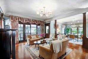 Un salon spacieux doté d’une grande fenêtre, d’un lustre et d’un coin repas avec des meubles recouverts d’un tissu à motifs floraux.