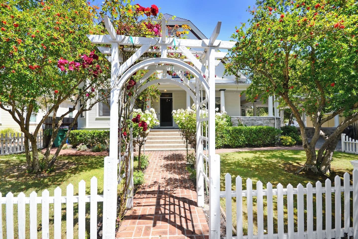 A white wooden archway leading to a house with a brick pathway.