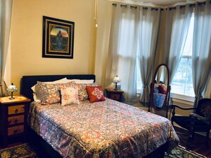 Une chambre à coucher avec un lit, des tables de chevet, une chaise, un miroir et une fenêtre avec des rideaux.