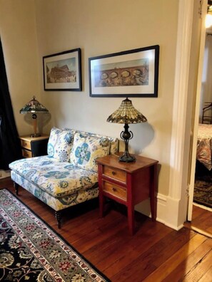 A room with a patterned sofa, a small wooden nightstand with a lamp, and two framed pictures on the wall. - Victorian 2 bedroom sleeps six by Forsyth Park (Savannah)