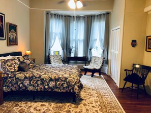 A bedroom with a bed, two chairs, a nightstand, a lamp, and a window with curtains. - Victorian 2 bedroom sleeps six by Forsyth Park (Savannah)