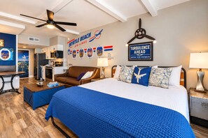 Une chambre d’hôtel équipée d’un lit, d’un bureau, d’un canapé, d’un ventilateur de plafond et d’un mur orné de logos de compagnies aériennes et d’un panneau d’avertissement concernant les turbulences.