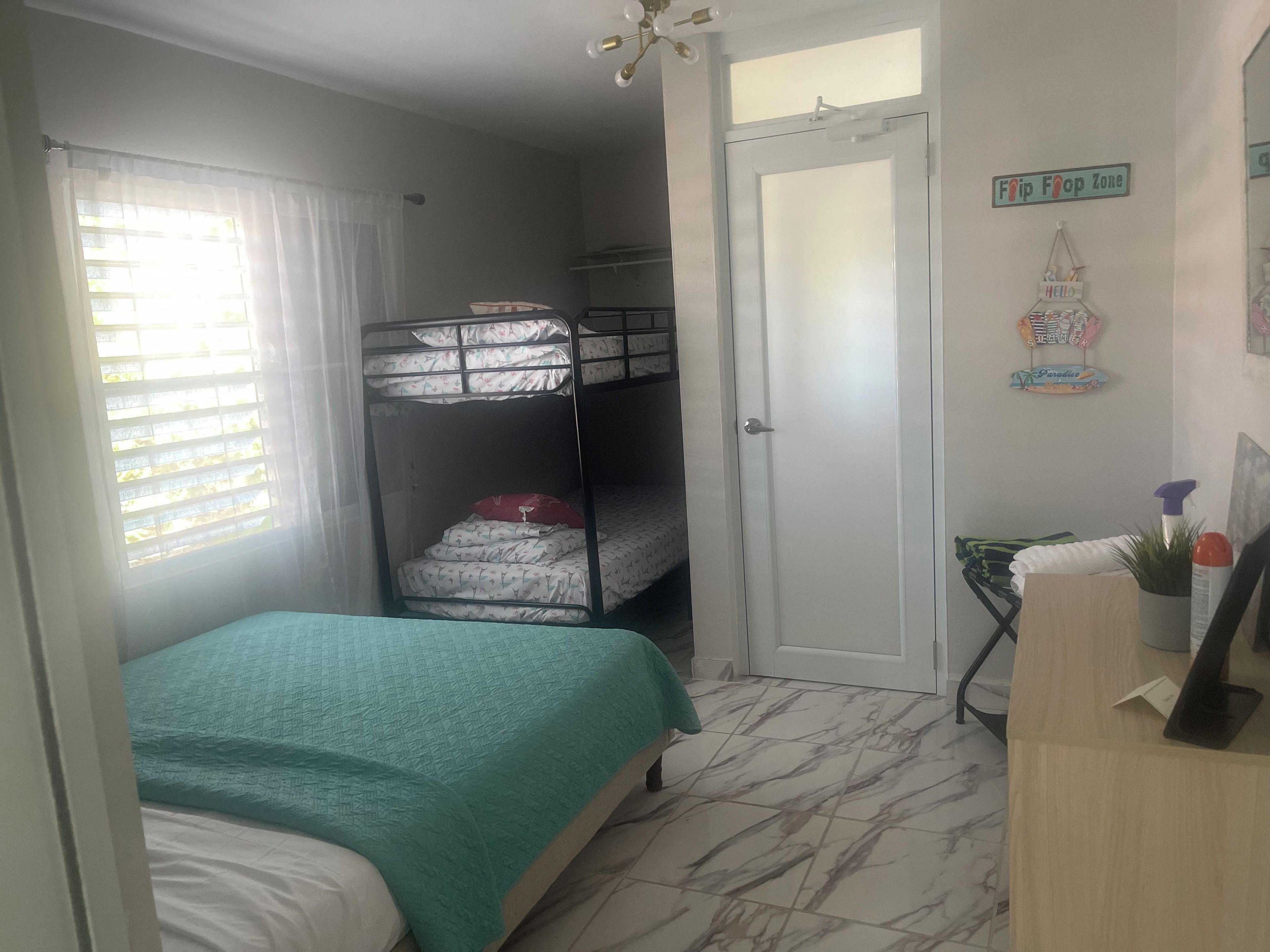 A bedroom with a bunk bed, a single bed, a desk with a chair, a door, and a window with curtains.