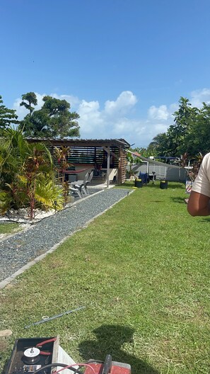 Uma área externa gramada com uma estrutura de madeira, possivelmente um deck ou varanda, e um pequeno jardim com diversas plantas.