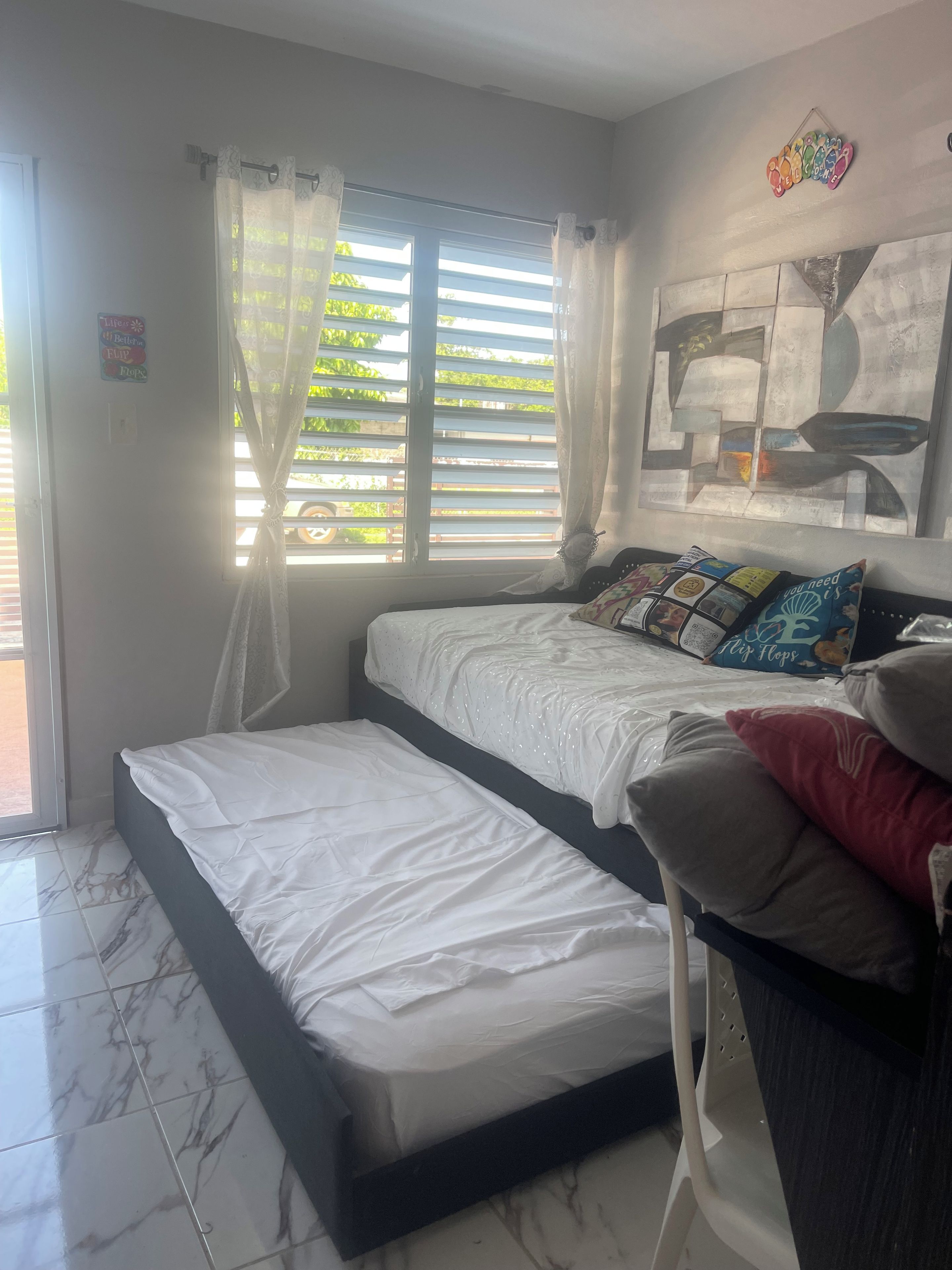 A bedroom with a bed, a window with curtains, a wall art piece, and a tiled floor.