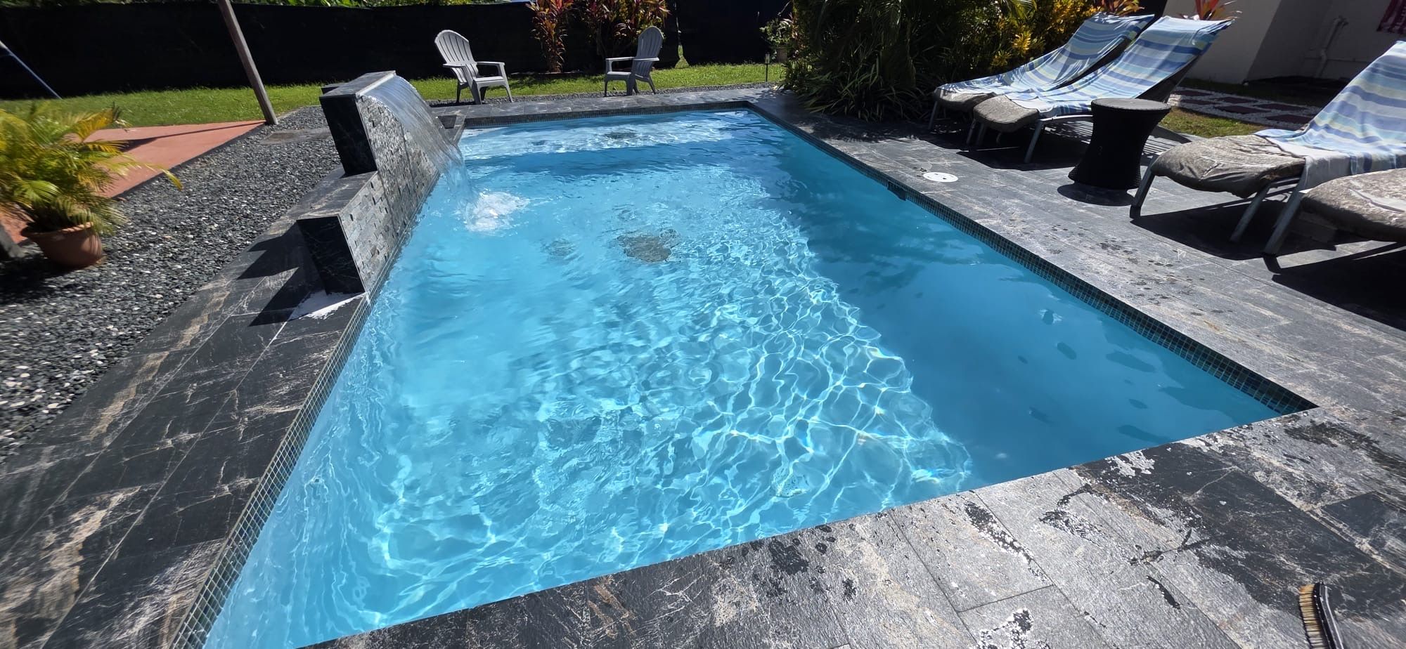 A rectangular swimming pool with a clear blue water surface, surrounded by a stone deck and equipped with lounge chairs.