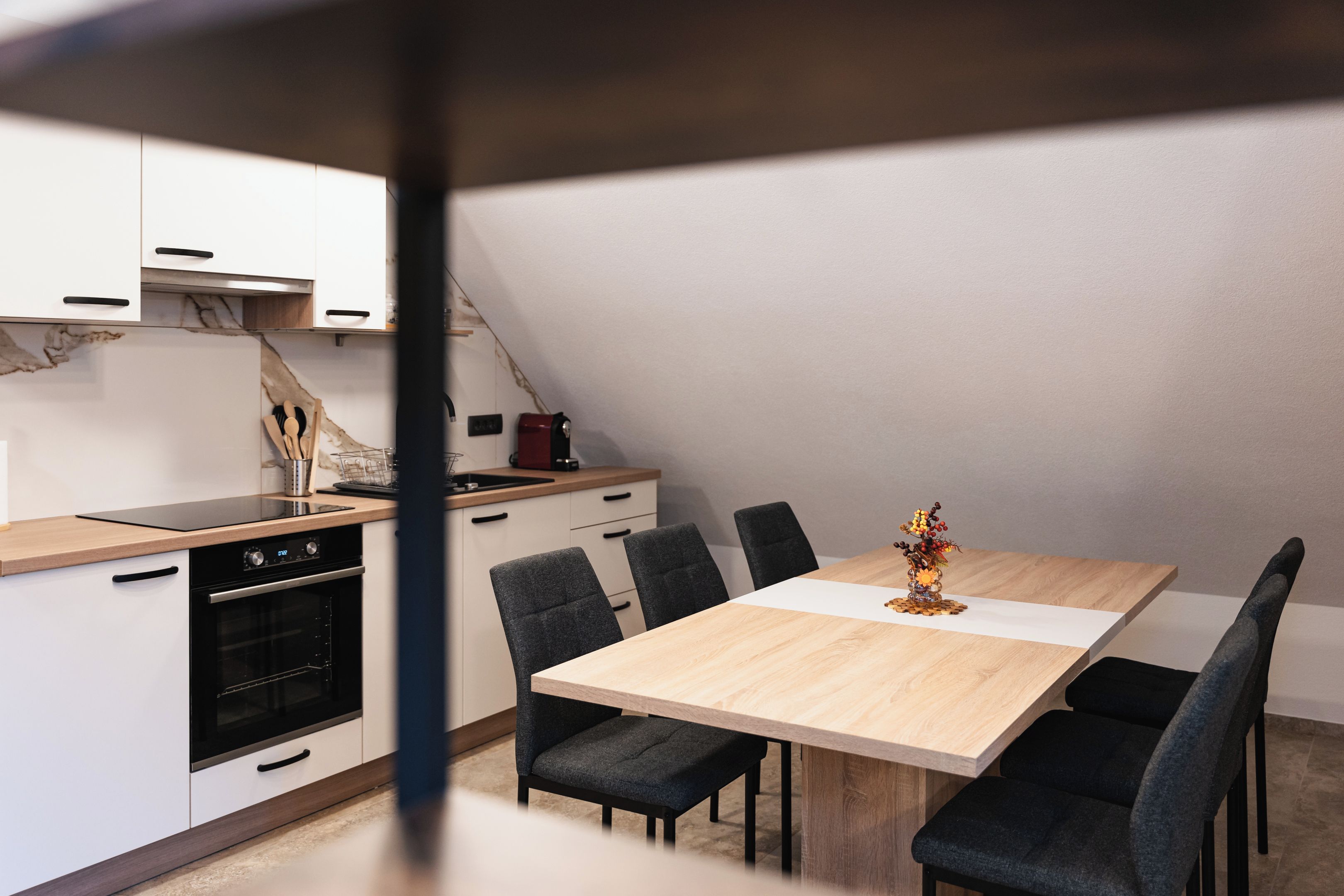 Een moderne keuken met een inbouwoven, een magnetron en een koffieapparaat. Er staat een eettafel met zes stoelen bij, en een vaas met bloemen erop.