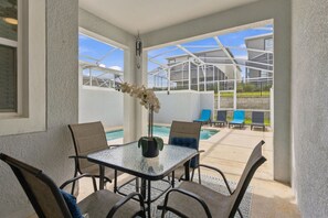 En täckt uteplats med ett glasbord, stolar, en krukväxt och utsikt över en swimmingpool och en byggnad.