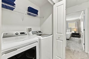 A laundry room with two white washing machines, a drying rack with folded towels, and a door leading to a bedroom with a bed and a nightstand. - Magical Getaway with Private Pool, Top location (Davenport)