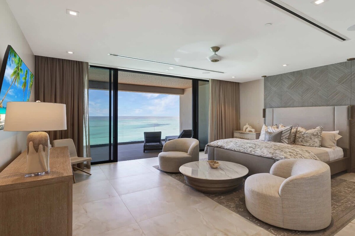 A modern hotel room with a large bed, a seating area with two chairs, a coffee table, a TV, and a view of the ocean.