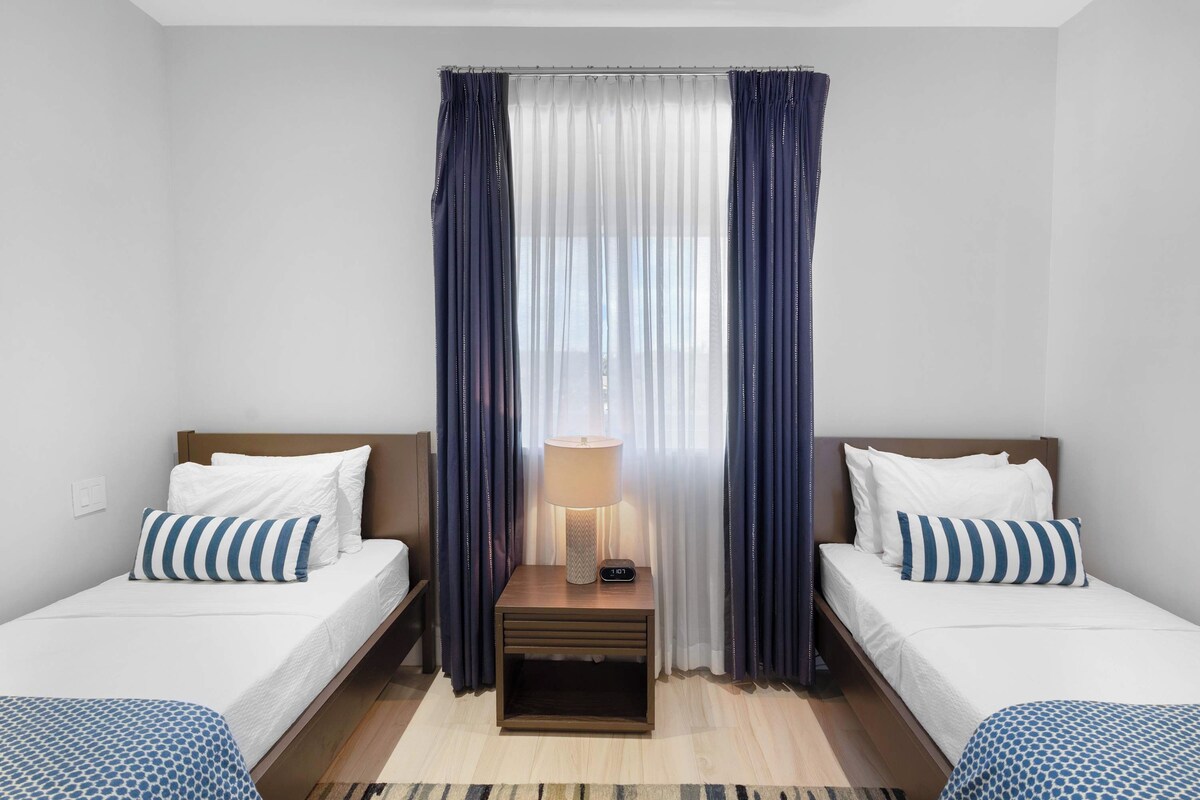 A hotel room with two single beds, a bedside table with a lamp, and a window with curtains.