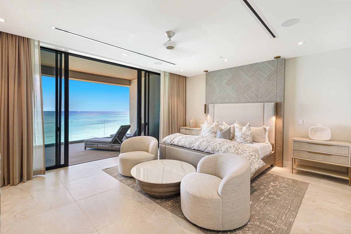 A modern bedroom with a large bed, two armchairs, a round coffee table, and a view of the ocean.