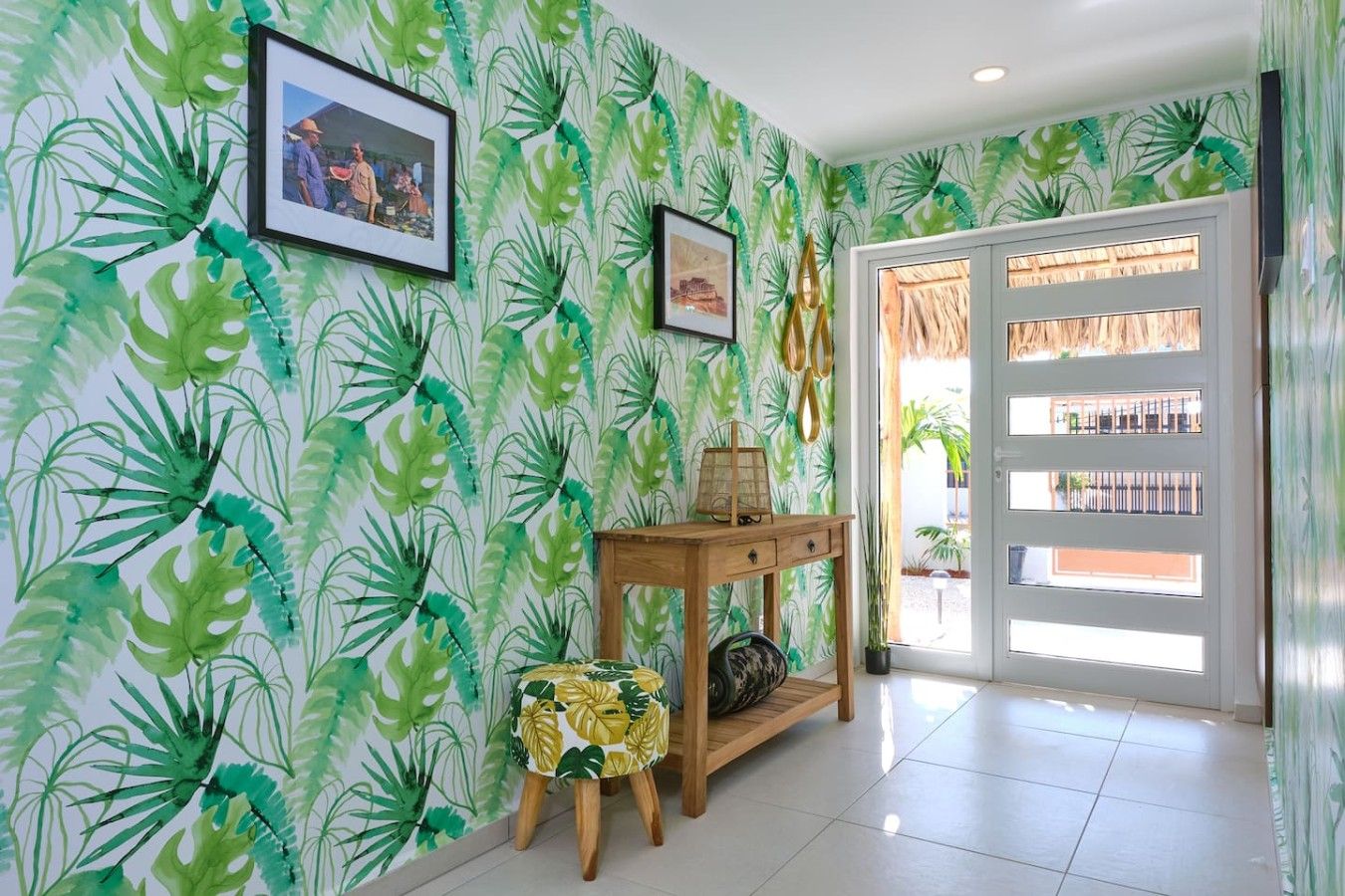 A room with a tropical leaf pattern wallpaper, a wooden console table with a lamp, and a small stool.