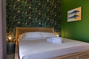 Ein ordentlich bezogenes Bett mit weißen Leinen und einem gefalteten Handtuch, ein hölzernes Kopfteil und eine Wand mit einem grünen Blattmuster.