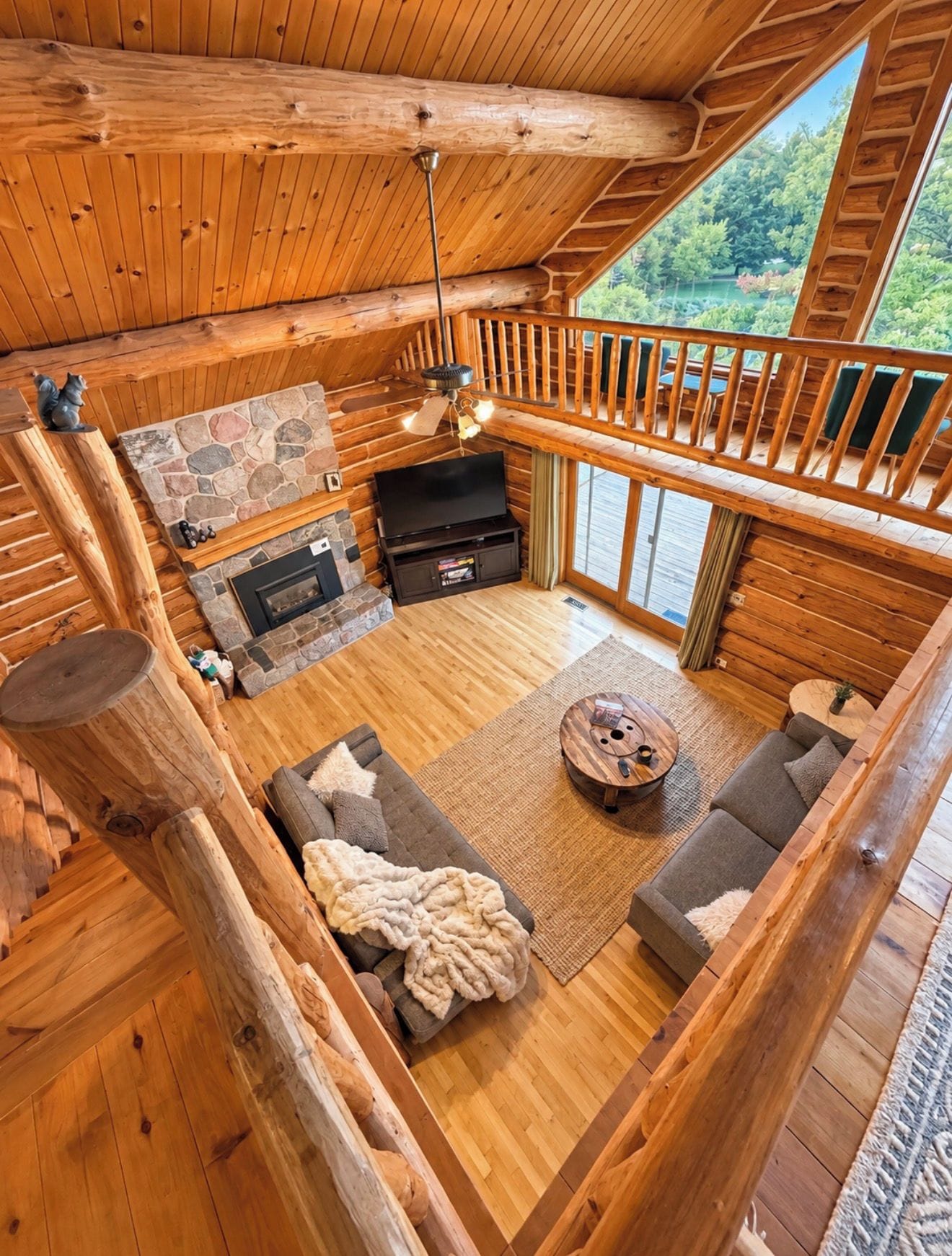 Ein Interieur einer Blockhütte mit Kamin aus Stein, einem Fernseher und einem Balkon mit Blick auf den Wald.