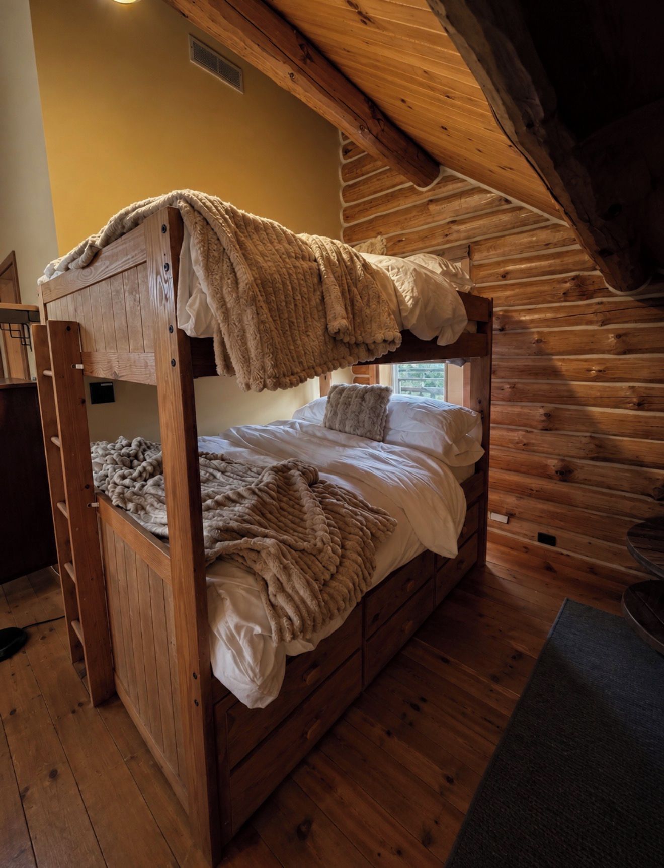 A bunk bed with a wooden frame and a cozy blanket, set in a room with wooden walls and a sloped ceiling.