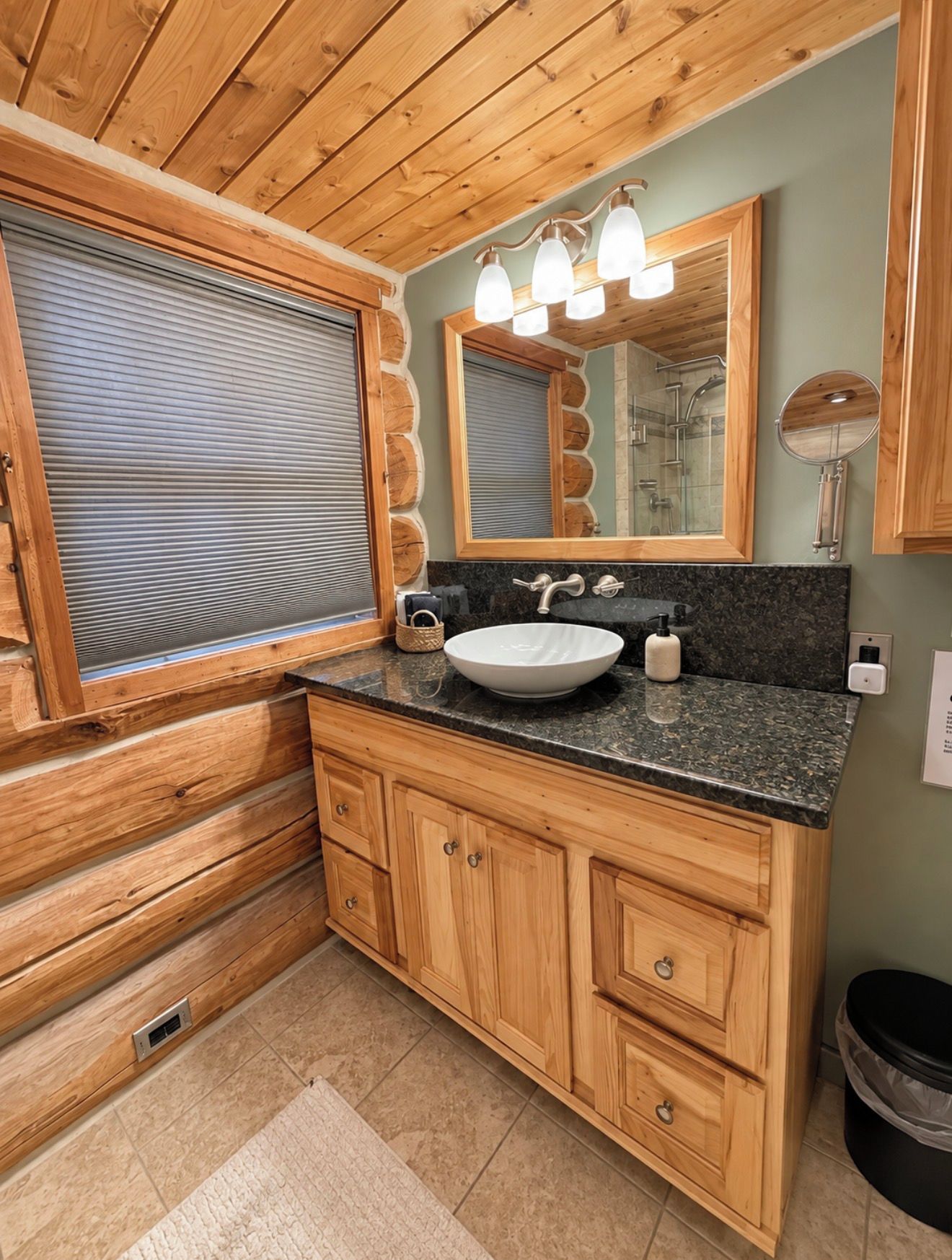 A wooden cabin-style bathroom with a large window, a sink with a mirror above, and wooden cabinets.