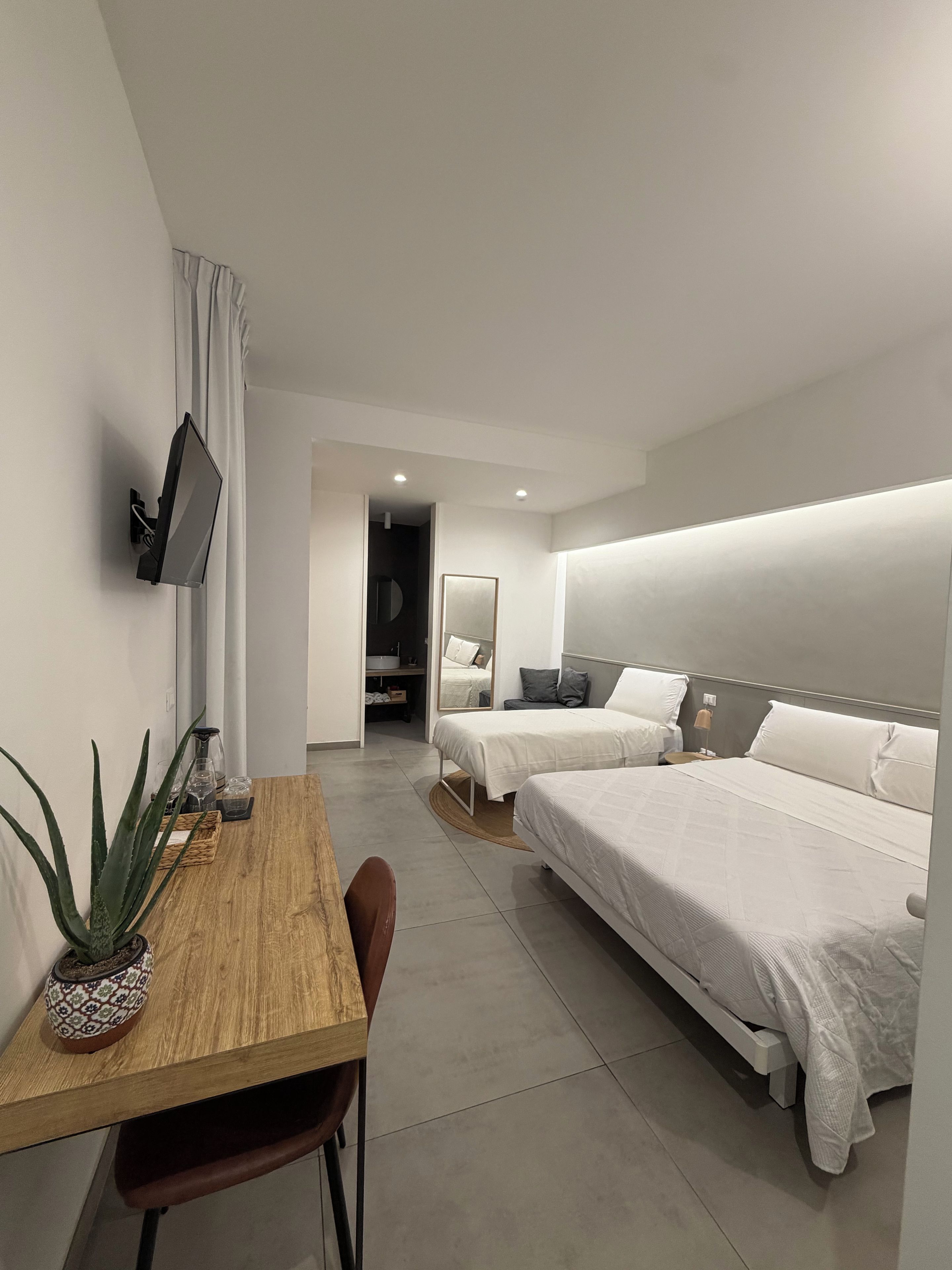 A modern hotel room with a wooden desk, two beds, a flat-screen TV, and a mirror.