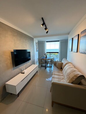A modern living room with a flat-screen TV, a sofa, a dining area with a table and chairs, and a balcony with a view. - Incredible Ocean View 2 Bedrooms | Itaparica Beach (Vila Velha)